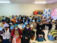 About 30 women sit in a classroom, wearing surgical masks. Each one olds a tablet computer and shows it to the camera.